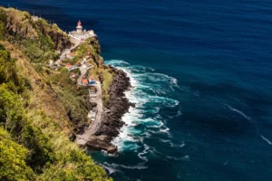 Vista aérea do Farol do Arnel no Nordeste, falésia verde, casas e mar azul profundo de São Miguel.